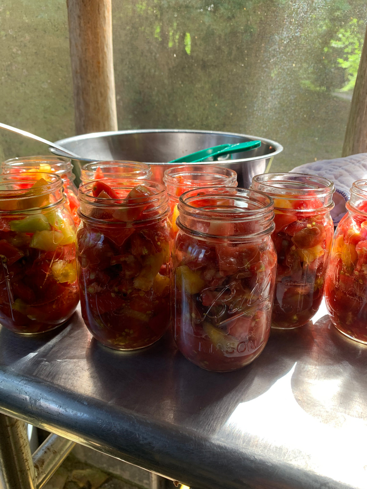 Several clear glass mason jars filled with heirloom tomatoes and herbs, arranged on a metal tray outdoors near a window.