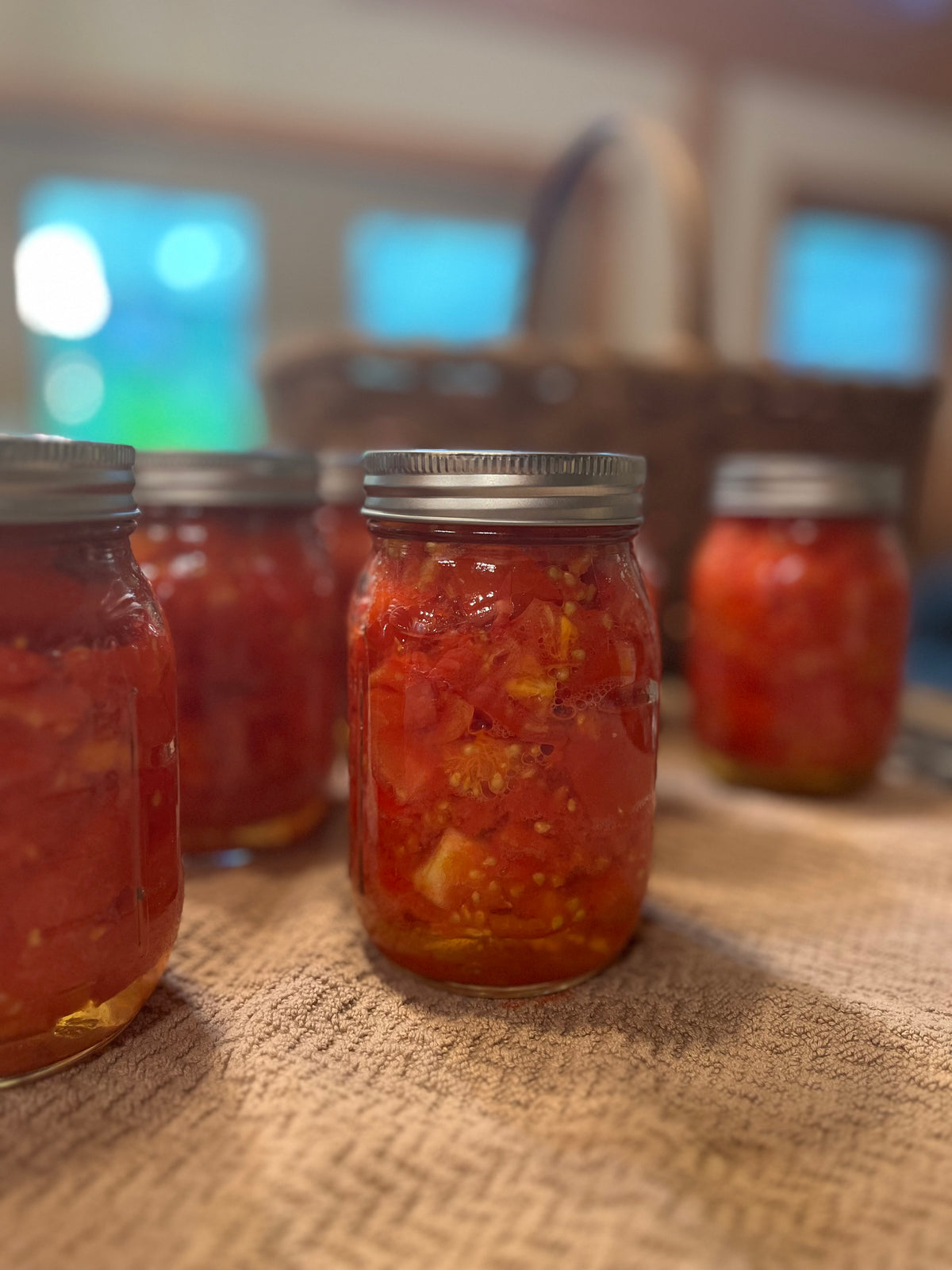 Several glass mason jars filled with chopped red tomatoes and other vegetables, sealed with silver metal lids, arranged on a woven tablecloth in an indoor setting with a blurred background.